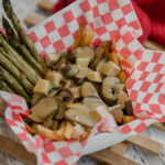 A loaded poutine with mushrooms and asparagus in basket lined with checkered paper.