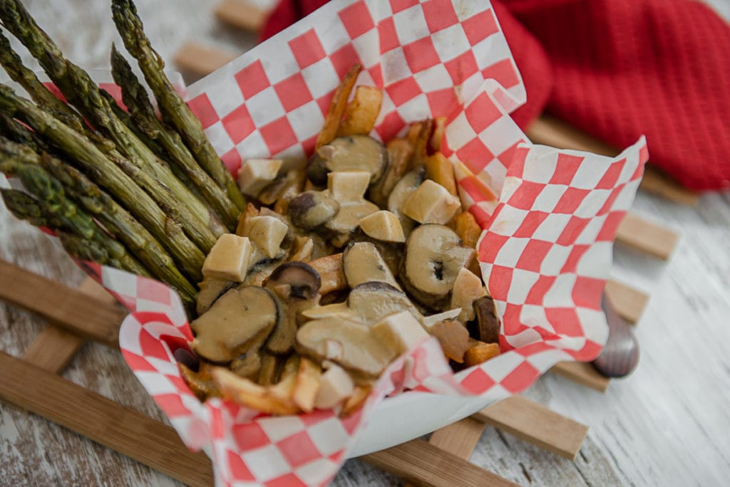 A loaded poutine with mushrooms and asparagus in basket lined with checkered paper.