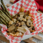 A hearty vegan poutine with mushroom, creamy gravy and roasted asparagus in a bowl lined with checkered red and white wax paper.