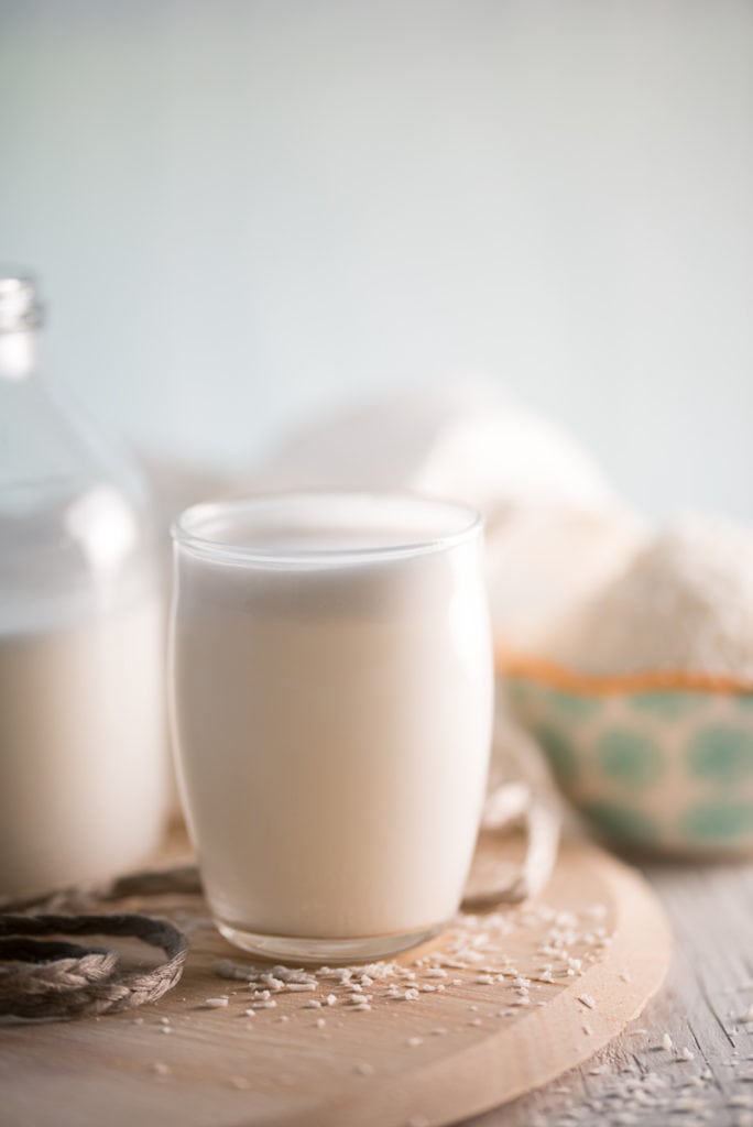A glass of coconut milk sitting in front of a half full jar and an overflowing bowl of shredded coconut.