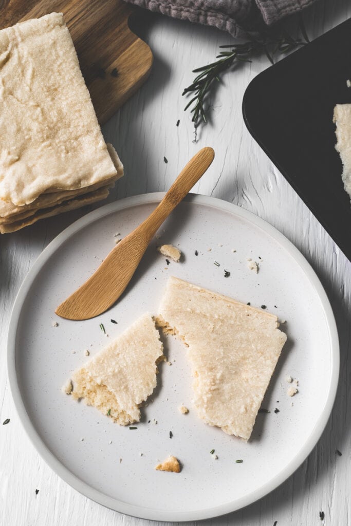 A half-eaten piece of flatbread on a plate surrounded by a stack of flatbread, a sprig of rosemary and a baking sheet.