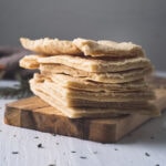 Homemade Flatbread stacked on a wooden slab with spilt herb crumbs and a linen napkin in the background.