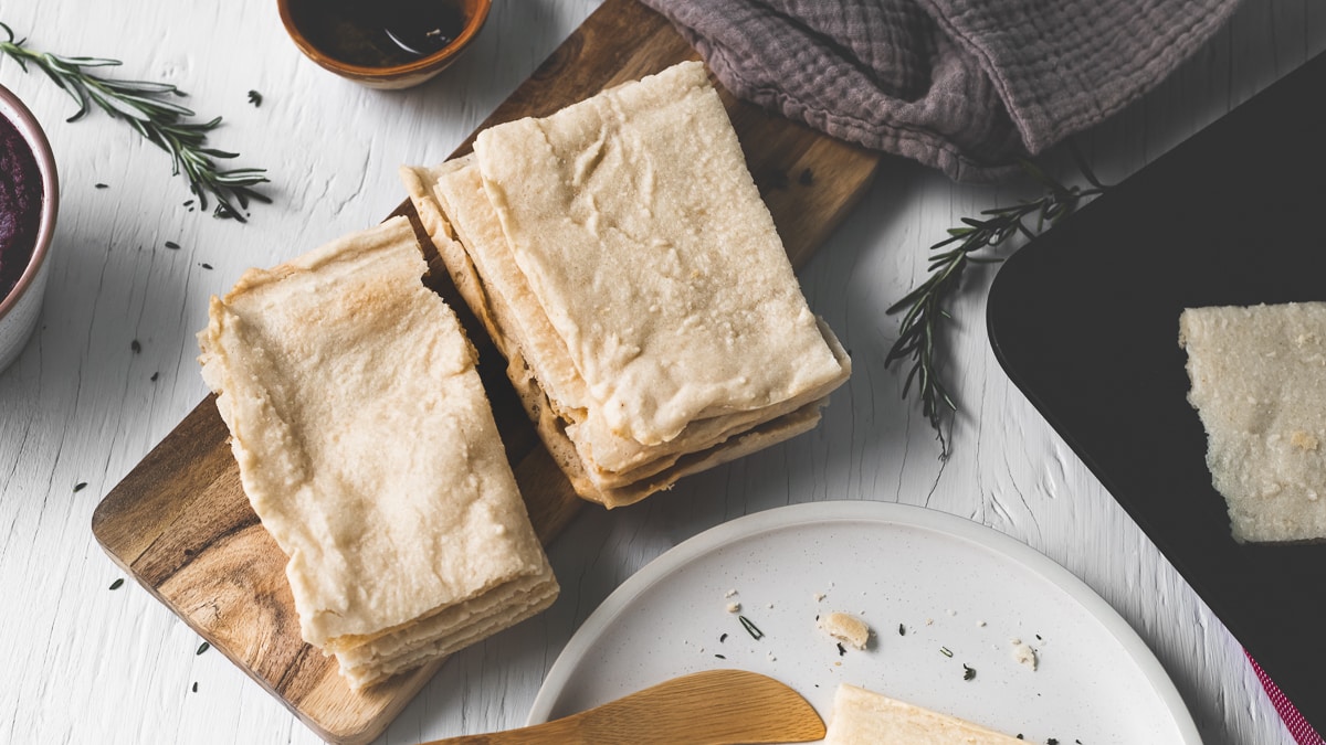 Two stacks of flatbread on a wooden slab beside a plate with a half-eaten piece, a linen cloth and a bowl of oil.