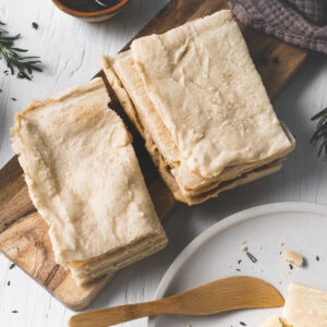 Two stacks of flatbread on a wooden slab beside a plate with crumbs and a butter knife, a linen cloth and a bowl of oil.