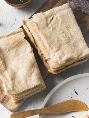 Two stacks of flatbread on a wooden slab beside a plate with crumbs and a butter knife, a linen cloth and a bowl of oil.