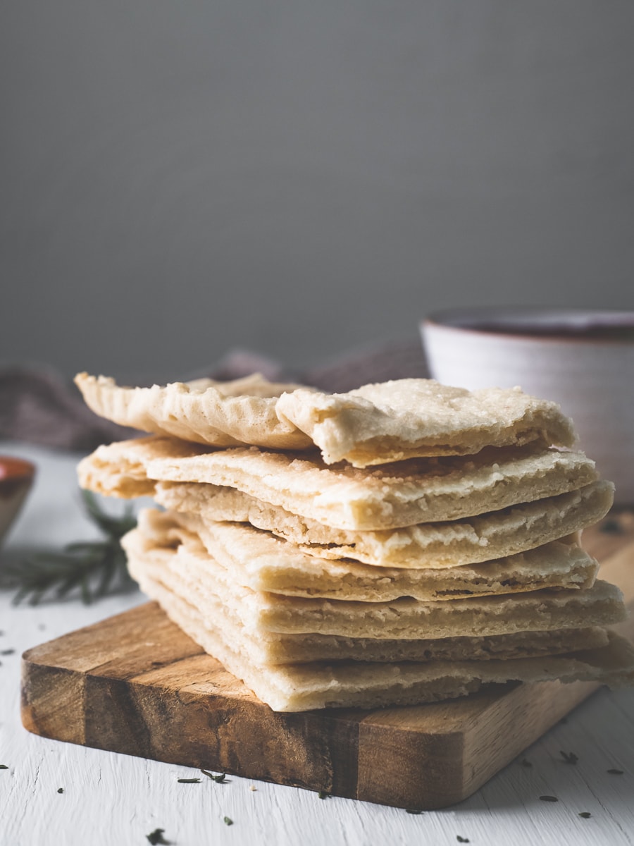 A stack of homemade flatbread on a wooden slab cutting board surrounded by crumbs, spilt herbs and bowls filled with bread dippers.