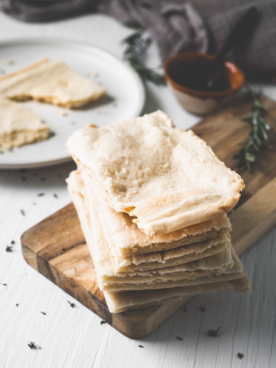 A stack of flatbread on a wooden slab in front of a plate and a bowl of oil.