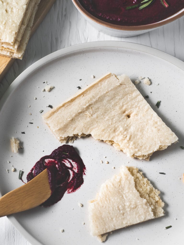 A half-eaten piece of flatbread on a plate full of crumbs, a swirl of beetroot hummus and a butter knife.