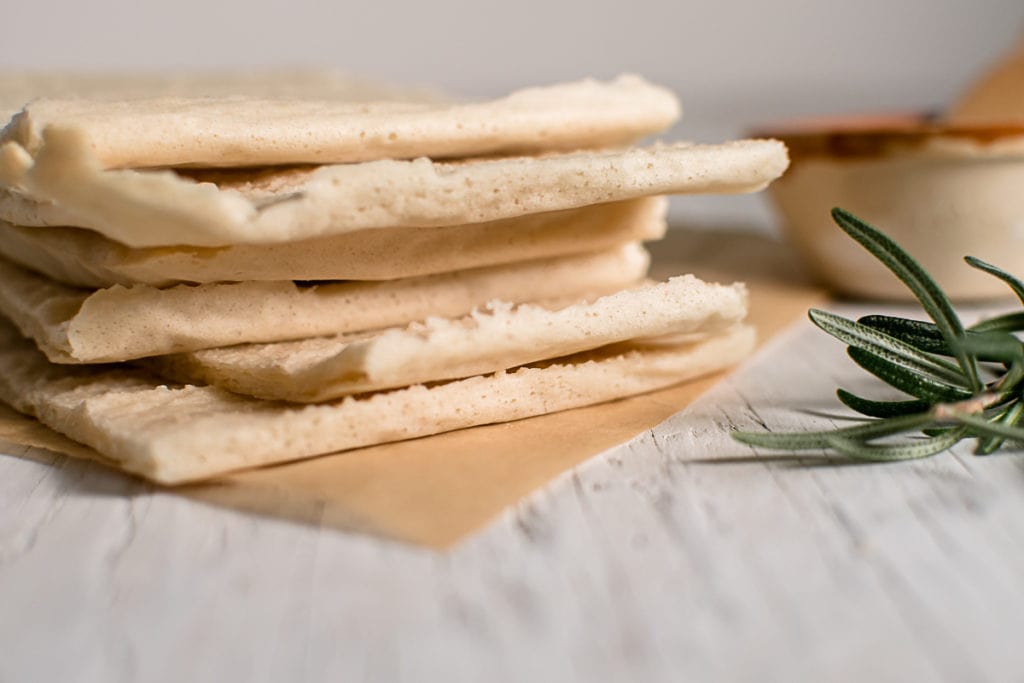 A stack of flatbread squares beside a sprig of rosemary and bowl of oil.