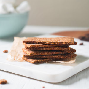 A stack of healthy homemade Graham Crackers on a crinkled piece of parchment on top of a marble cutting board. Behind the stack of Graham Crackers is a bowl of marshmallows and surrounding is chocolate crumbs.