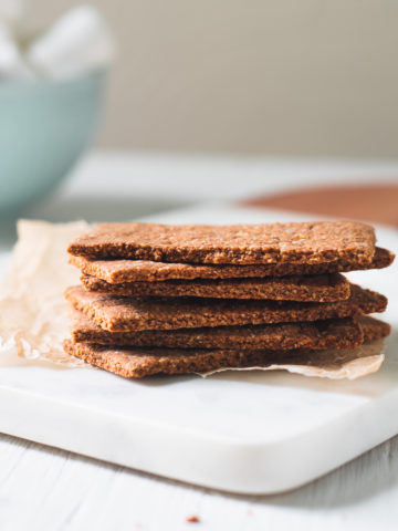 A stack of healthy homemade Graham Crackers on a crinkled piece of parchment on top of a marble cutting board. Behind the stack of Graham Crackers is a bowl of marshmallows and surrounding is chocolate crumbs.