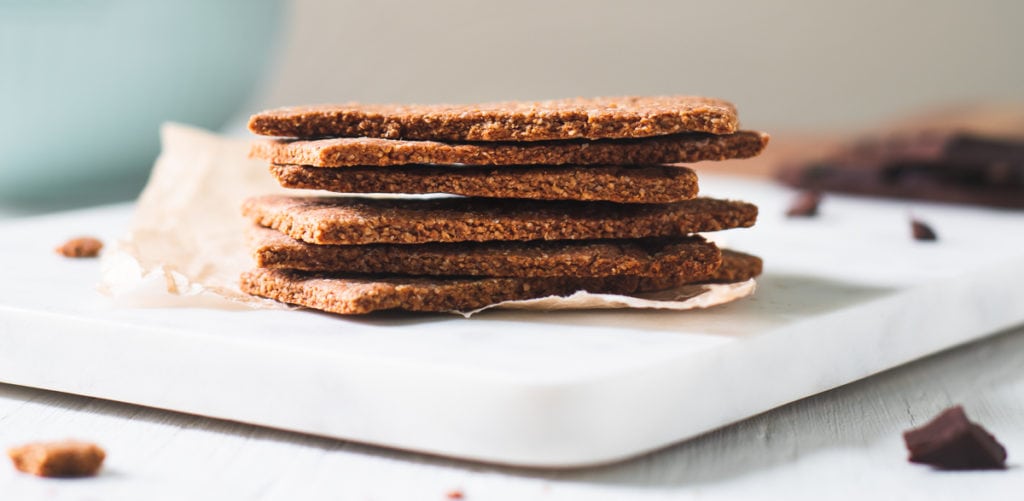 A stack of Homemade Graham Crackers on a crinkled piece of parchment on top of a marble cutting board. Surrounding the Graham Cracker stack is chocolate crumbs and a teal bowl.