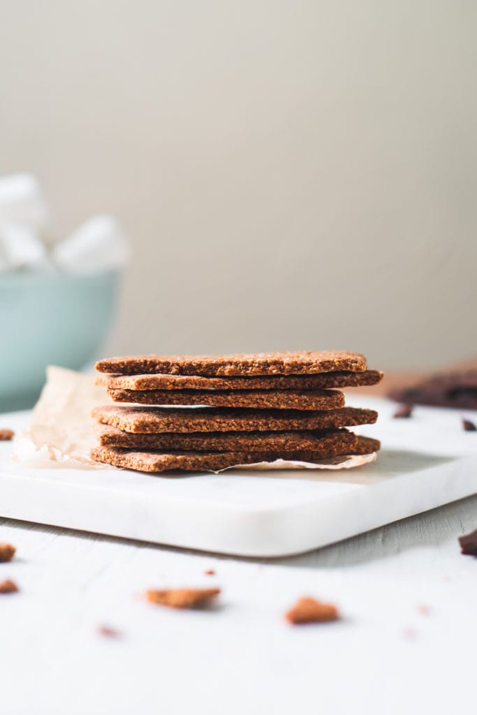 A stack of homemade graham crackers sitting on top of a marble cutting board with a bowl of marshmallows and chocolate and cookie crumbs surrounding.