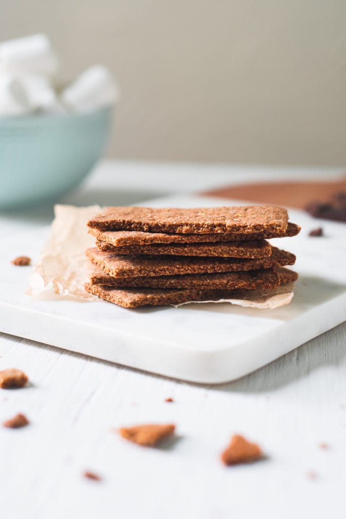 A stack of homemade gluten-free Graham Crackers on a crinkled piece of parchment on top of a marble cutting board. Behind the stack of Graham Crackers is a bowl of marshmallows and surrounding is chocolate crumbs.