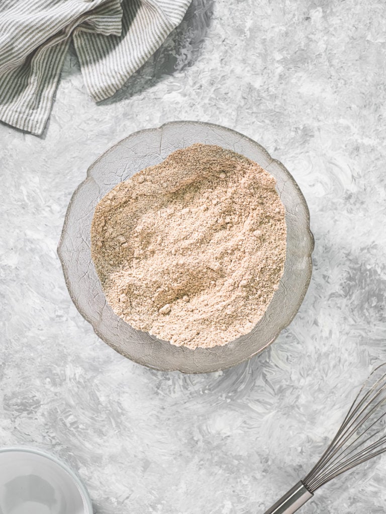 A mixing bowl of flour with a bowl and a whisk below and a striped cloth above.