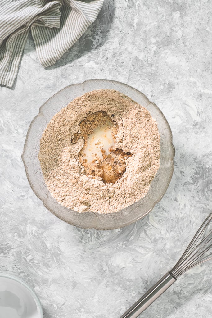 A mixing bowl of flour, non-dairy milk and coconut oil with a whisk above and a linen cloth above.