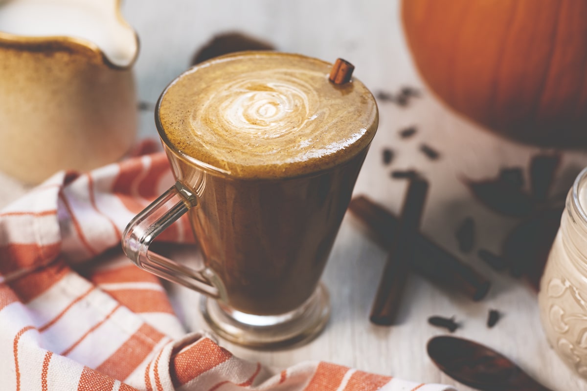 A richly coloured pumpkin spice latte swirled with cashew cream in a glass mug beside a pumpkin, cinnamon sticks, creamer jug and orange napkin.