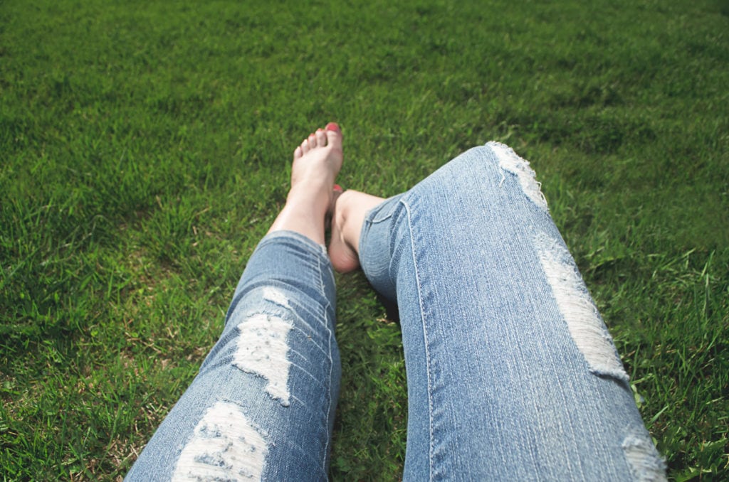 The legs of a woman wearing jeans resting barefoot in the grass.