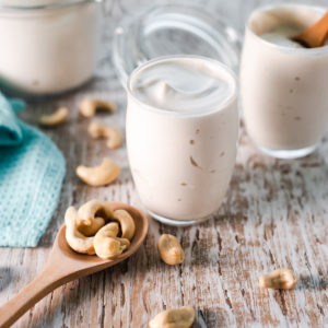 Cashew cream served into glass cups from a latch jar with a spoonful of spilled cashews in front.