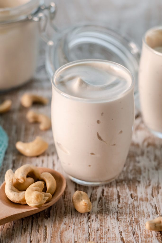 Creamy cashew yogurt swirled in a cup centred in between a spoonful of spilled cashews and a latch jar.