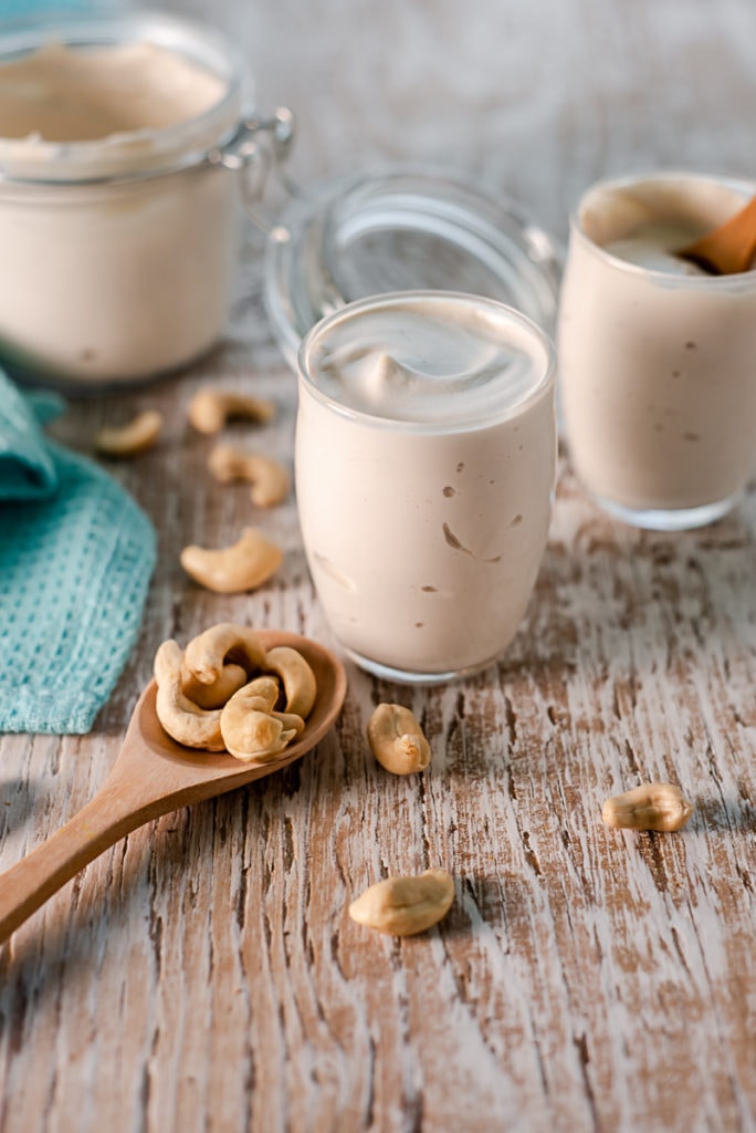 Cashew cream served into glass cups from a latch jar with a spoonful of spilled cashews in front.