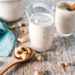 A little cup of creamy cashew yogurt, swirled to the top with another cup full and a larger jar behind, as well as a spoon of cashews spilling about.