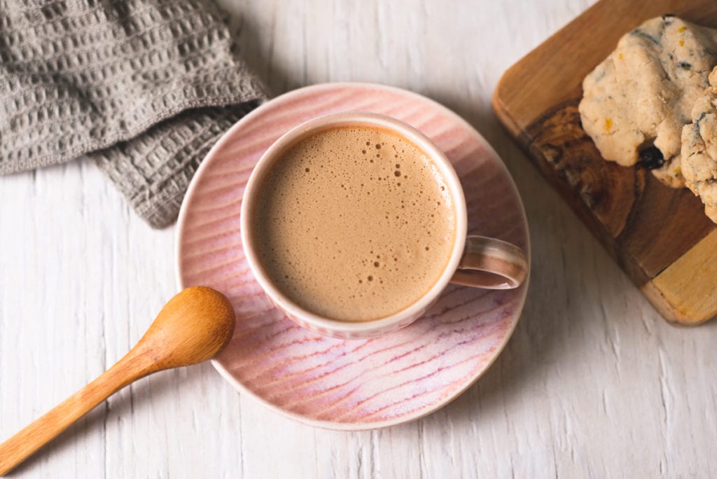 A frothy dandelion latte in a pink espresso cup on a matching plate with a wooden spoon resting on the edge.