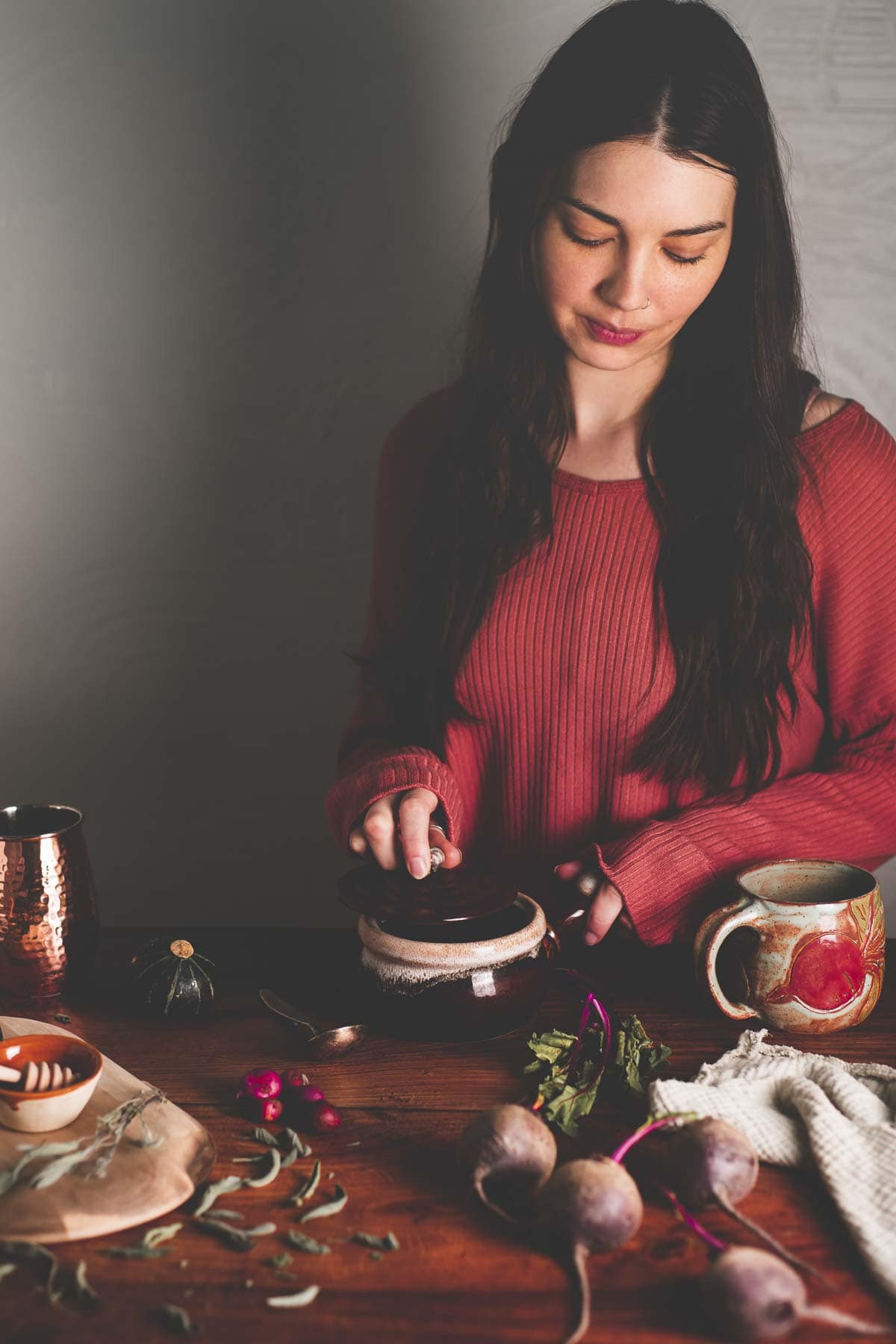 A dark-haired woman preparing whole foods on a rustic kitchen table with fresh root vegetables.