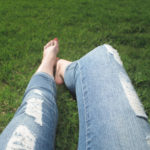 The legs of a woman wearing jeans resting barefoot in the grass.