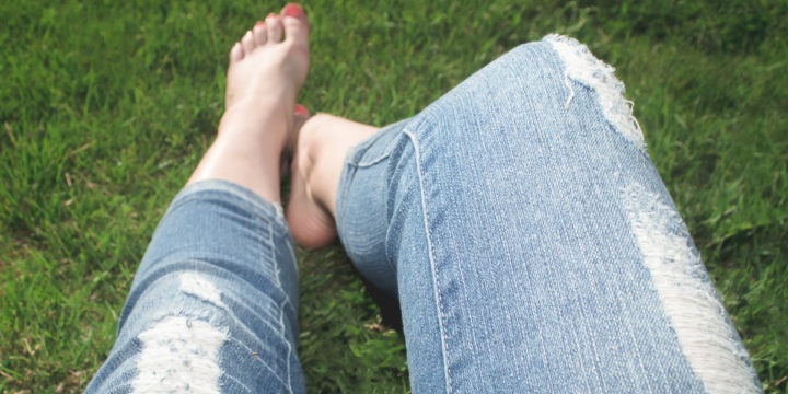 The legs of a woman wearing jeans resting barefoot in the grass.