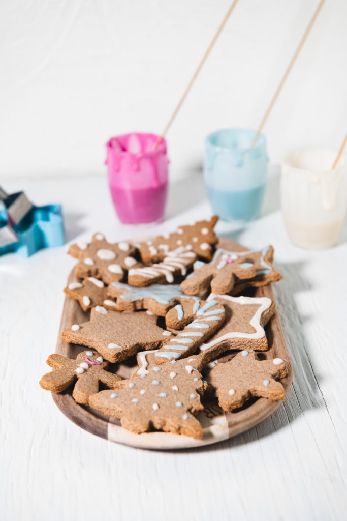A plate full of blue and white decorated gingerbread cookies surrounded by cups of eggless royal icing and cookie cutters