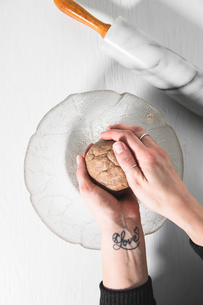 Hands rolling gluten-free gingerbread dough into a ball over a glass mixing bowl and a rolling pin to side.