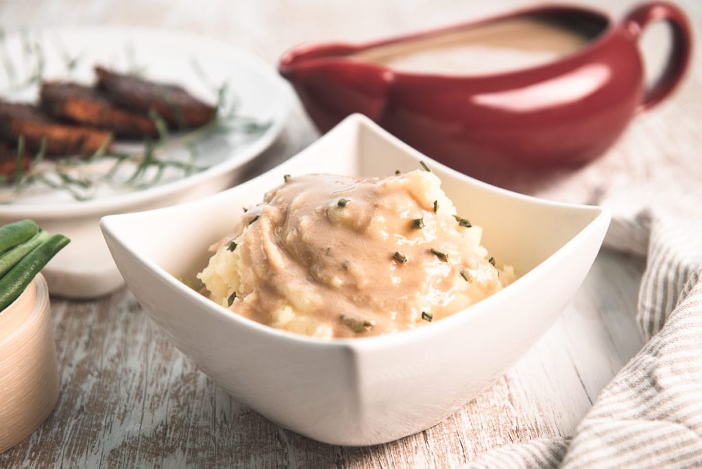 A gravy-soaked bowl of mashed potatoes in front of a gravy boat and tempeh slices.