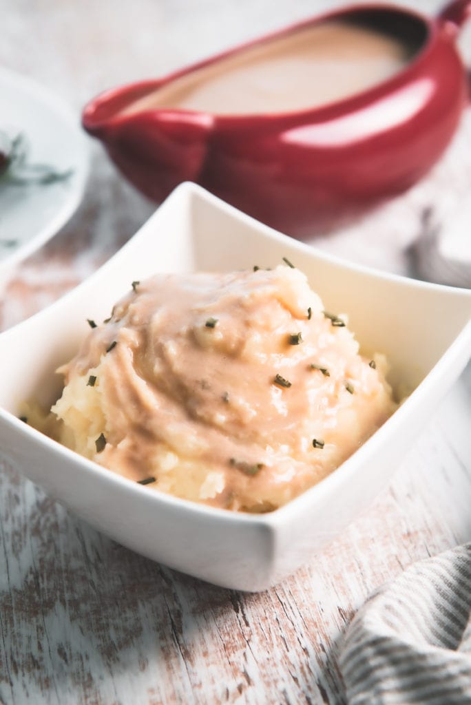 A gravy-soaked bowl of mashed potatoes in front of a gravy boat and tempeh slices.