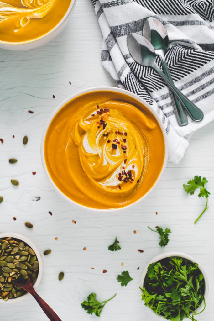 Two hearty bowls of roasted butternut squash soup swirled with cream and topped with crushed pepper flakes. Surrounding the bowls are toasted pumpkin seeds, fresh sprigs of parsley and a linen napkin holding two spoons, ready to dig in.