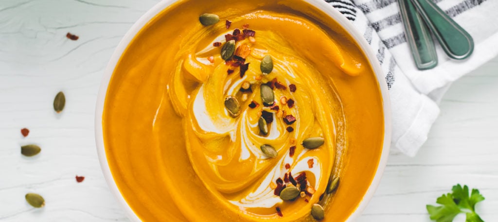 A bowl of Butternut Squash Soup swirled with cashew cream and topped with crushed pepper flakes and toasted pumpkin seeds. Surrounding the bowl is fresh sprigs of parsley, spilled pumpkin seeds and pepper flakes and two spoons crossed on a linen napkin.
