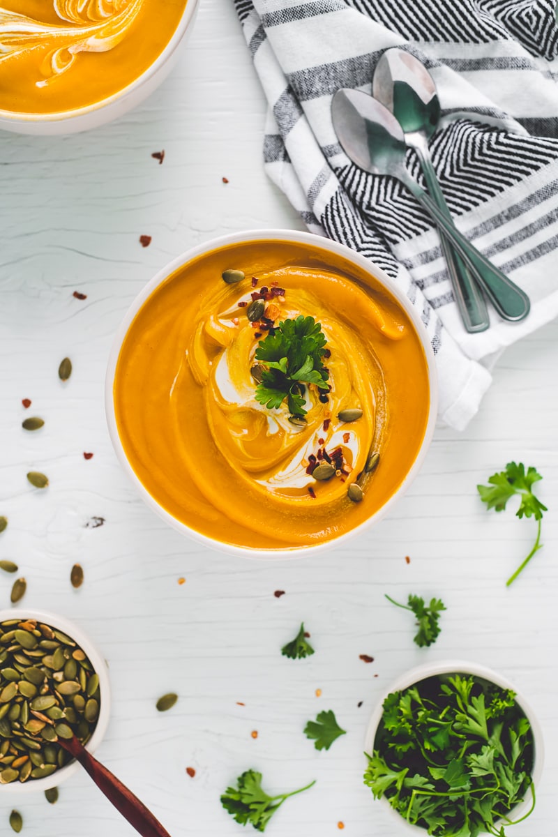 Two large bowls of vibrant butternut squash soup swirled with cream and garnished with toasted pumpkin seeds, crushed pepper flake and a few sprigs of fresh parsley.