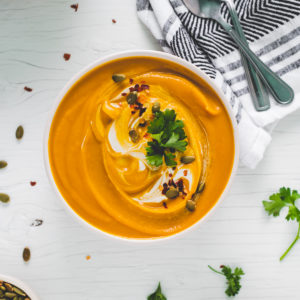 A big bowl of vibrant butternut squash soup swirled with cream and garnished with toasted pumpkin seeds, crushed pepper flake and a few sprigs of fresh parsley.