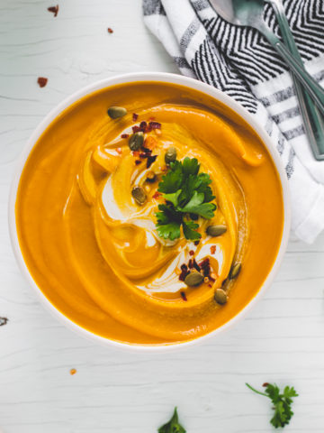 A big bowl of vibrant butternut squash soup swirled with cream and garnished with toasted pumpkin seeds, crushed pepper flake and a few sprigs of fresh parsley.