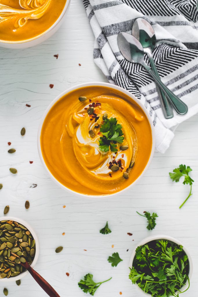 Two large bowls of vibrant butternut squash soup swirled with cream and garnished with toasted pumpkin seeds, crushed pepper flake and a few sprigs of fresh parsley. A bowl of pumpkin seeds and bowl of parsley sit below with some spilt all around. A napkin with two spoons sits above, ready to be enjoyed.