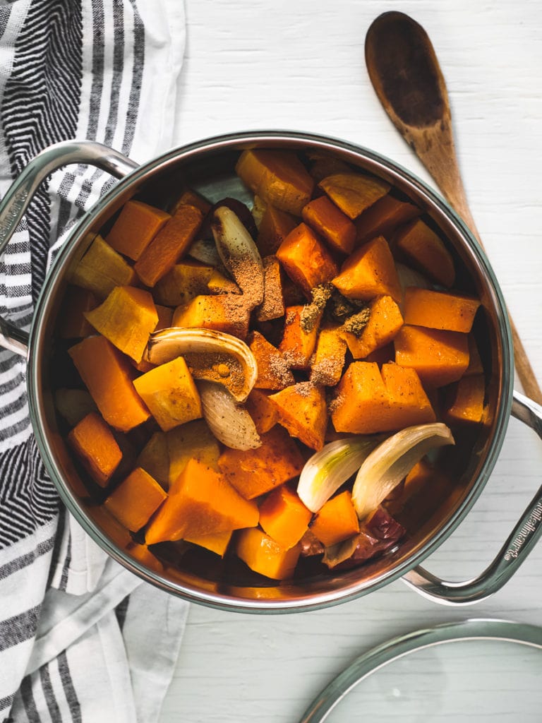 A large soup pot half-way filled with roasted butternut squash, onion, apple and garlic. A striped linen napkin and rustic wooden spoon surround the pot.