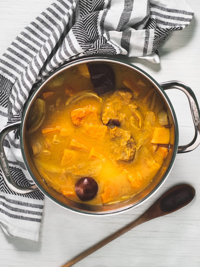 A large soup pot filled with roasted butternut squash, onion, apple and garlic simmering in a mixture of vegetable broth and dairy-free milk. Above is a striped linen napkin cradling the pot and a rustic spoon lays below.
