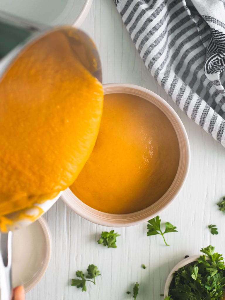 Steamy butternut squash soup being poured into a large soup bowl with fresh sprigs of parsley dropped around and a linen napkin scrunched to the side.