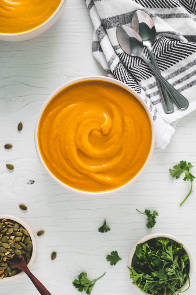 Thick, hearty and vibrant butternut squash soup swirled in two bowls with pumpkin seeds, fresh parsley ready for garnishing and two spoons laying crossed on a striped linen napkin.