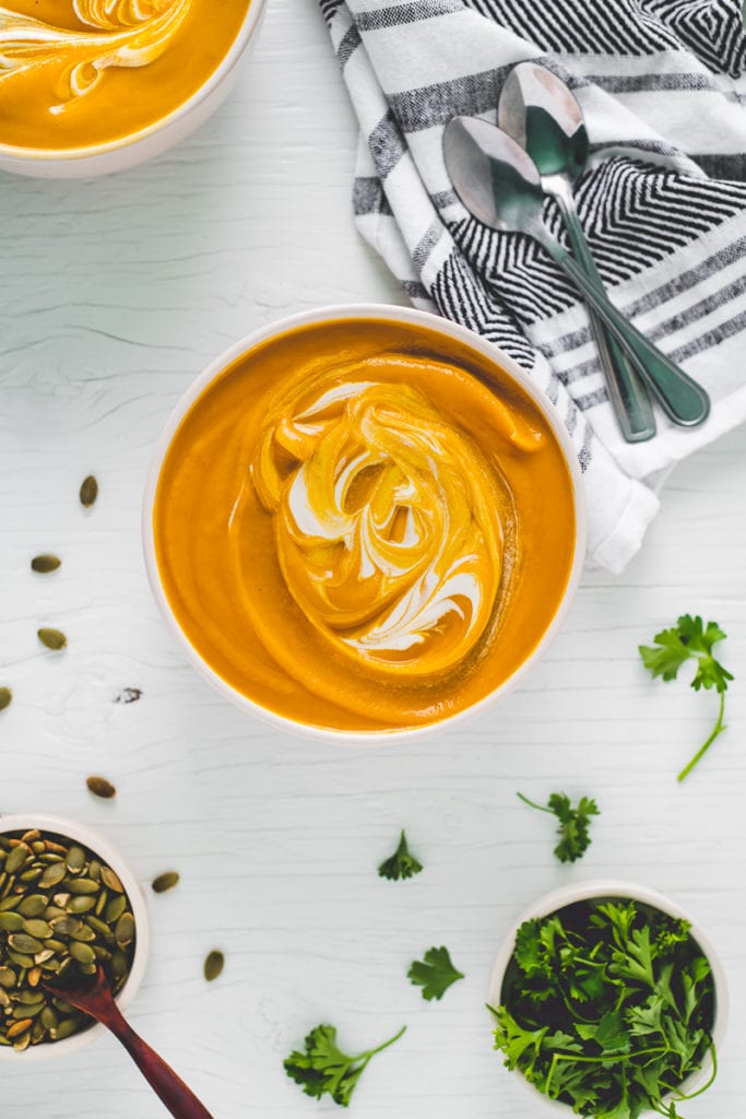 Two large bowls of roasted butternut squash soup swirled with cashew cream and surrounded by toasted pumpkin seeds and fresh parsley for garnishing.