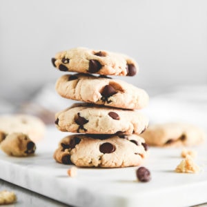 A stack of golden-brown chocolate chip cookies surrounded by crumbs.