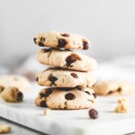 A stack of golden-brown chocolate chip cookies surrounded by crumbs and chocolate chips.