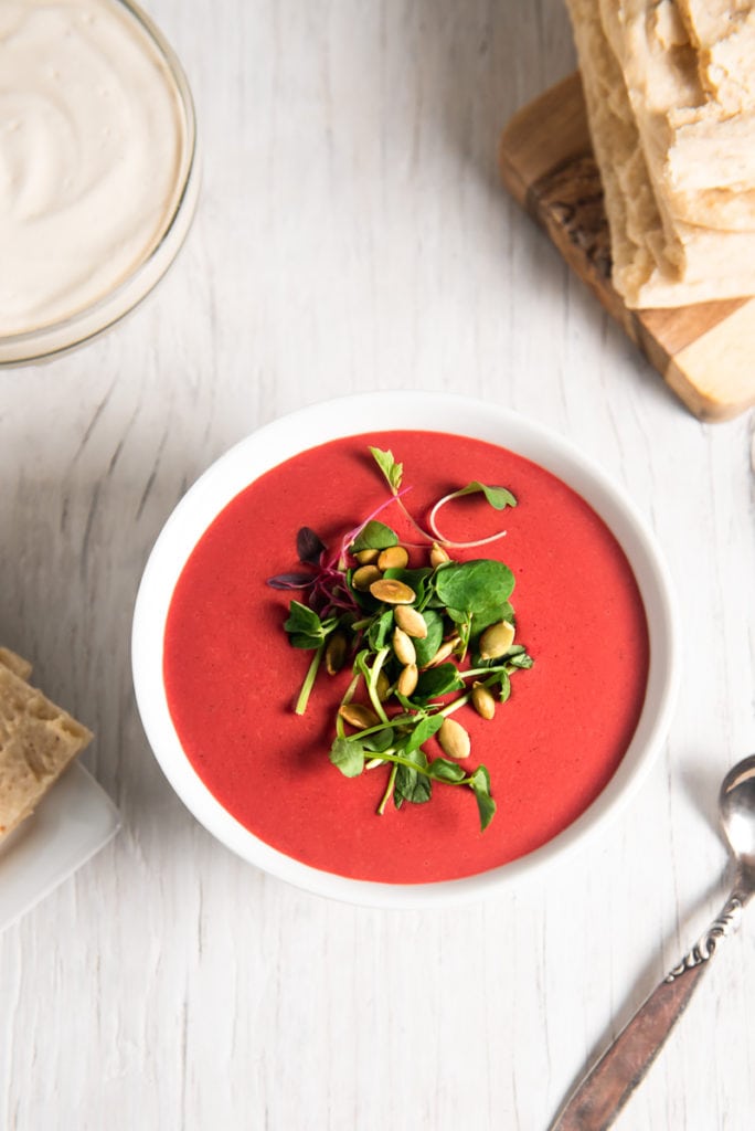A bowl of beet soup ready to be eaten with a spoon, flatbread, sprouts and cream.