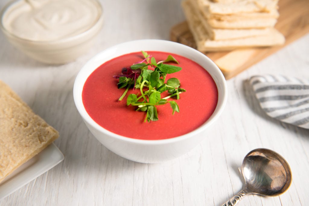 A bowl of vibrant, creamy beetroot soup garnished with fresh sprouts and pumpkin seeds in front of a stack of flatbread and bowl of cashew cream.