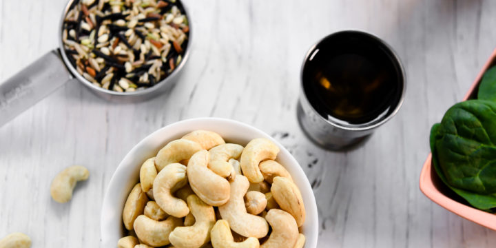 A little bowl of cashews with a few spilled on the table along with a bowl of olives, a measuring cup of wild rice, a little cup of olive oil and a bunch of bananas.